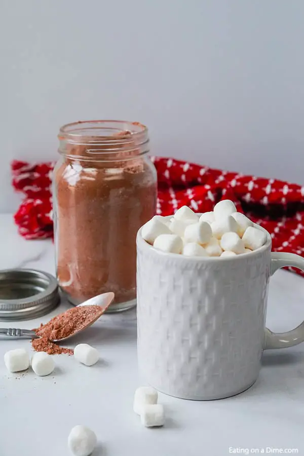 Close up image of hot chocolate mix in a mason jar with a spoon. Also a white mug with hot chocolate with marshmallows. 