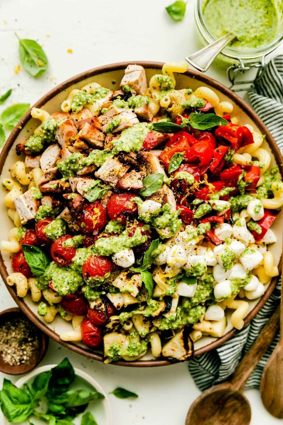 An overhead shot of a large bowl of prepared pasta salad topped with pesto, sitting alongside a wooden cutting board, bowl of basil leaves, striped dish towel, jar of pesto, and bowl of pepper atop a white surface.