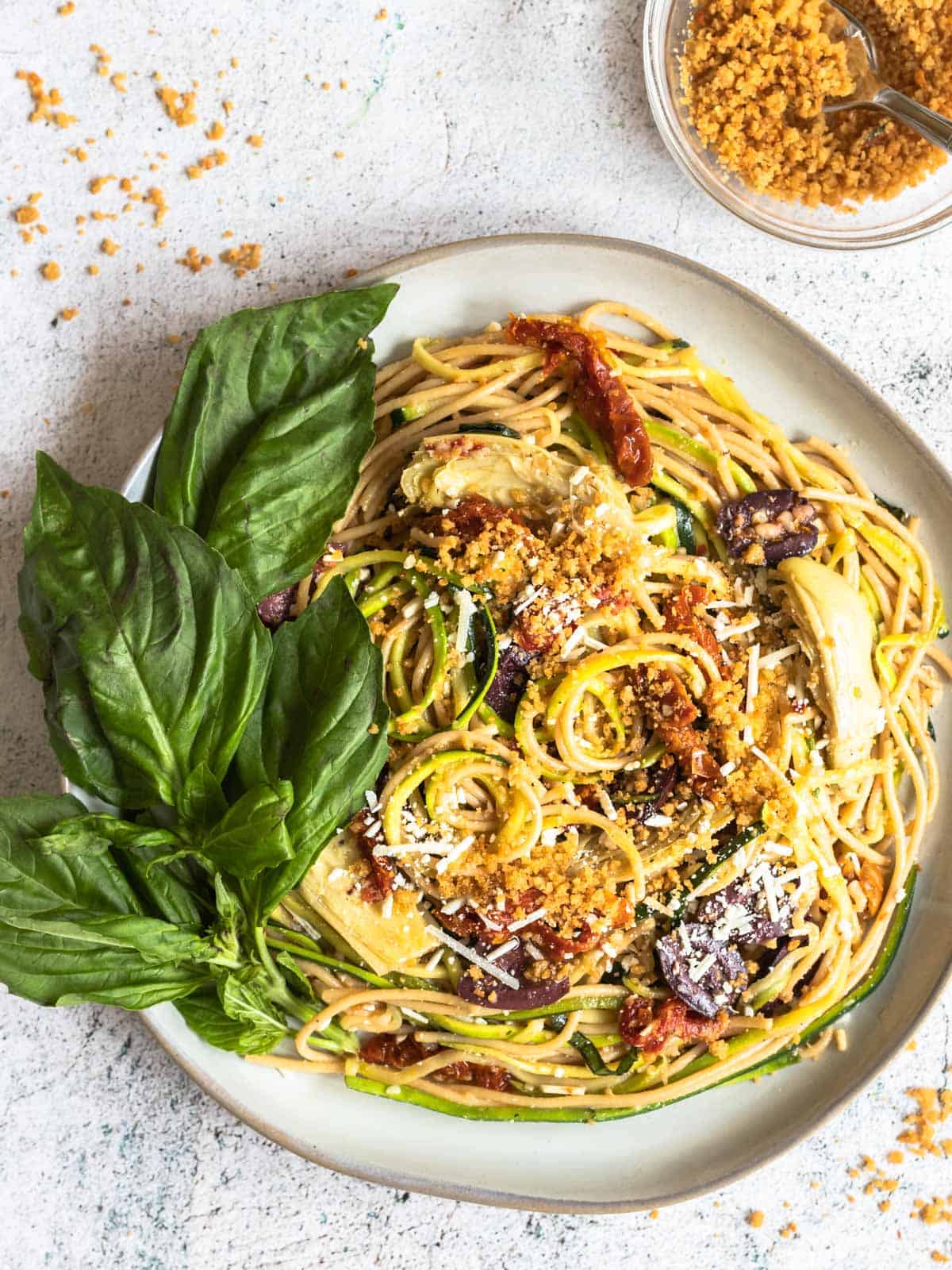 Mediterranean zucchini spaghetti on a plate next to a bowl of toasted breadcrumbs with a spoon